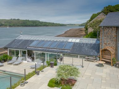 A patio with lounge chairs and plants overlooking a water body with a stone building and glass roof extension at Curlew House in Cosheston