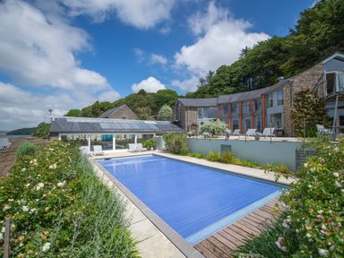An outdoor swimming pool covered with a blue pool cover next to a stone building with large windows and patio chairs at Curlew House in Cosheston