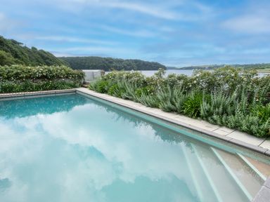 An outdoor swimming pool with surrounding plants and a view of trees and water at Curlew House in Cosheston