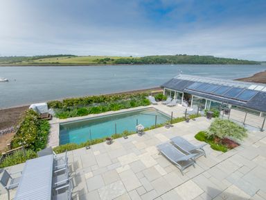 A patio with lounge chairs and dining table overlooking a swimming pool and a river at Curlew House in Cosheston