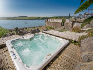 An outdoor hot tub on a wooden deck overlooking a river with a boat and grassy hills at Curlew House in Cosheston