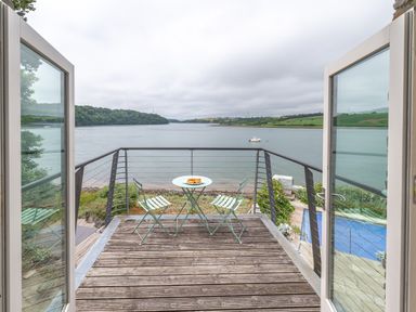 A balcony with a round table and three chairs overlooking a river and green hills at Curlew House in Cosheston