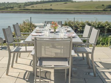 An outdoor dining table with eight chairs set with plates glasses and flowers by a river at Curlew House in Cosheston
