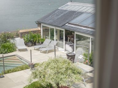 An outdoor patio with lounge chairs potted plants a pool and glass doors leading into a house at Curlew House in Cosheston