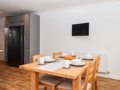 A dining area with a wooden table set for four with white dishes and mugs a black refrigerator and a wall-mounted television in the background at West View Northlew near Hatherleigh