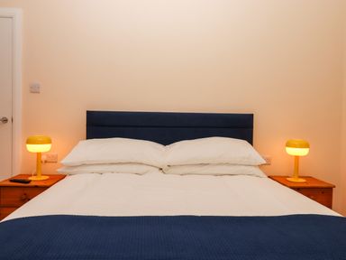 A bedroom with a double bed and blue headboard two bedside tables with yellow lamps and white walls at West View in Northlew near Hatherleigh