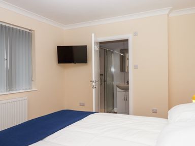 A bedroom with a bed white bedding a blue blanket a wall mounted TV a window with blinds and an open door leading to a bathroom with a shower at West View in Northlew near Hatherleigh