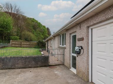 The exterior of a house with a white garage door a glass entrance door with a charging station mounted on the wall a small metal gate and a grassy yard surrounded by trees at West View Northlew near Hatherleigh