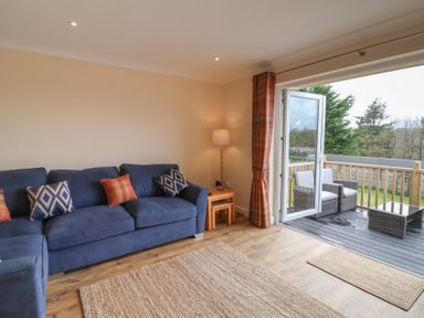 A living room with a blue sofa and cushions a floor lamp wooden side tables and an open door leading to an outdoor patio with wicker chairs at West View in Northlew near Hatherleigh