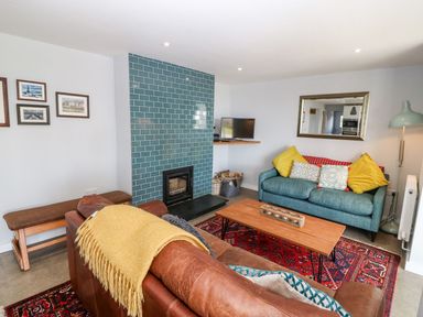 A living room with a brown leather sofa with a yellow throw a blue sofa with yellow and patterned cushions a wooden coffee table a red patterned rug a blue tiled fireplace a small TV on a corner shelf and framed photos on a white wall at Forest Lodge in Newborough