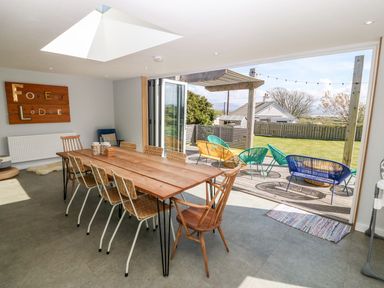 A dining room with a wooden table and chairs next to an open door leading to a garden with outdoor seating at Forest Lodge in Newborough