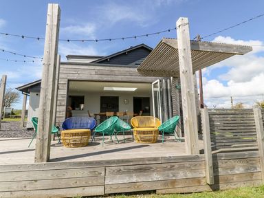A wooden deck with colorful outdoor chairs and a pergola connected to a house with open folding doors at Forest Lodge in Newborough