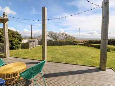 A deck with green chairs and a yellow table overlooking a fenced garden with a shed and trees at Forest Lodge in Newborough