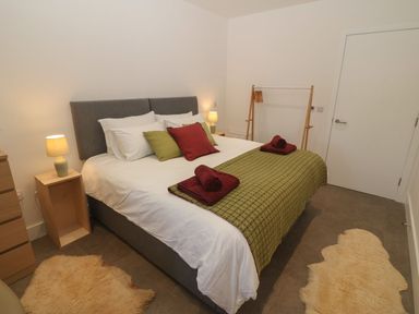 A bedroom with a double bed green and red cushions bedside tables with lamps a chest of drawers and a clothes rack at Tithe Barn in Capel Mawr near Malltraeth
