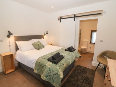 A bedroom with a bed and bedside tables and a bathroom visible through a sliding door at Tithe Barn in Capel Mawr near Malltraeth