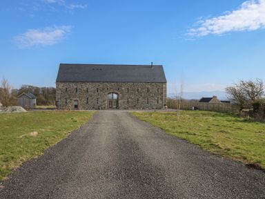 A gravel driveway leading to a stone barn with a pitched roof in a rural area at Tithe Barn in Capel Mawr near Malltraeth