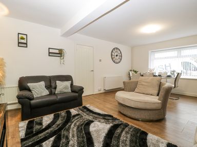 A living room with a black sofa two cushions a round beige armchair a dining table and chairs a wall clock and two small wall shelves at Meillion in Hirwaun