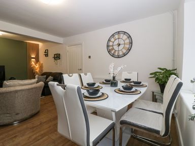 A dining area with a white table set with black dishes and six white and black chairs next to a living room with sofas and a wall clock at Meillion in Hirwaun