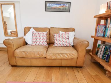 A room with a tan leather sofa with four pillows two with red and grey polka dots a wooden bookshelf with books and a framed landscape photo above the sofa at The Barn - Poplar Farm in Lower Dunsforth near Boroughbridge