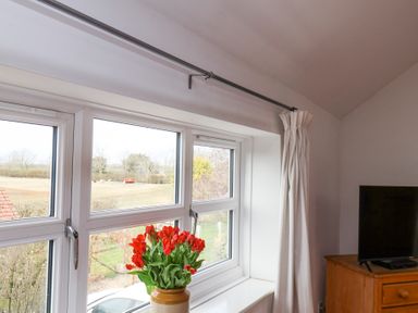 A window with red flowers in a vase on the sill and white curtains beside a wooden dresser with a TV on top at The Barn - Poplar Farm in Lower Dunsforth near Boroughbridge