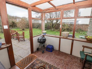 An enclosed porch area with a barbecue grill blue stool potted plants and a wooden chair looking out to a garden with patio furniture at Glan Yr Afon in Ystradgynlais