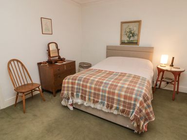 A bedroom with a bed covered with a plaid blanket a wooden chair a chest of drawers with a mirror and a side table with a lamp at Glan Yr Afon in Ystradgynlais