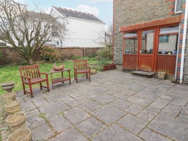 An outdoor patio area with stone tiles wooden chairs and a table near a brick house at Glan Yr Afon in Ystradgynlais