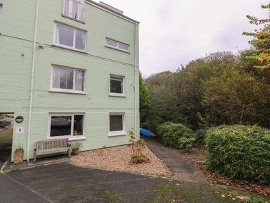 A green three story building with white framed windows a wooden bench and plants on a gravel area next to a paved walkway surrounded by trees and bushes at Kittiwake Apartment 22 in Little Haven near Broad Haven