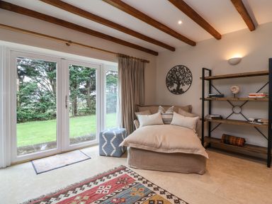 A living room with a large cushioned chair pillows a cube stool a wooden shelf with books and decor a rug and glass doors looking out to a garden at Blossom Hill near Witney