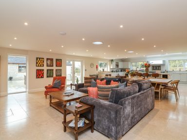 An open plan living room and kitchen with a large gray sofa and orange cushions with a wooden coffee table and dining table at Blossom Hill near Witney