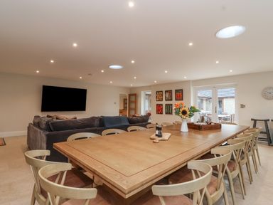 A dining area with a large wooden table surrounded by chairs and a living area with a dark sofa and wall-mounted TV at Blossom Hill near Witney
