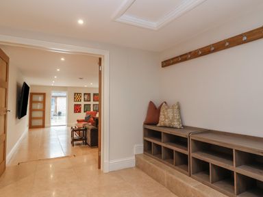 A hallway with wooden storage cubbies and hooks on the wall leading to a living area with a mounted TV and seating at Blossom Hill near Witney