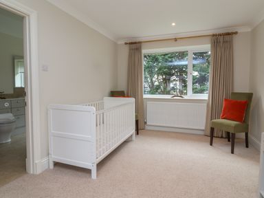 A nursery room with a white crib two green chairs with red cushions and a window showing trees at Blossom Hill near Witney