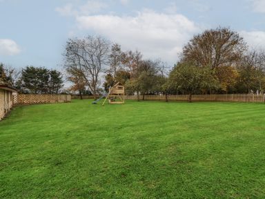 A large grassy backyard with a wooden play structure and slide surrounded by trees and wooden fences at Blossom Hill near Witney