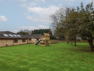 A grassy backyard with a play structure and trees near a stone building at Blossom Hill near Witney