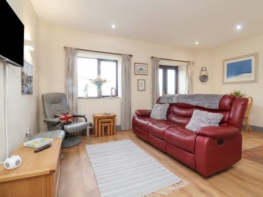 A living room with a red sofa gray armchair wooden tables and wall clock at Badgers Sett in Stibb near Kilkhampton