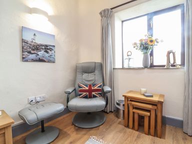 A corner with a gray chair and matching footrest with a union jack pillow a set of nested wooden tables and a window with flowers and lanterns at Badgers Sett in Stibb near Kilkhampton