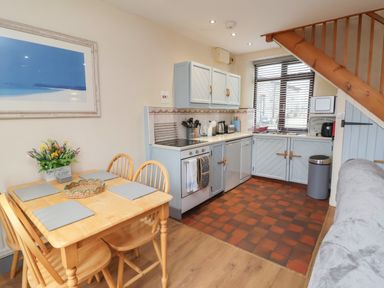 A kitchen and dining area with a wooden table and chairs a grey cabinet set under wooden stairs and a window with blinds at Badgers Sett in Stibb near Kilkhampton