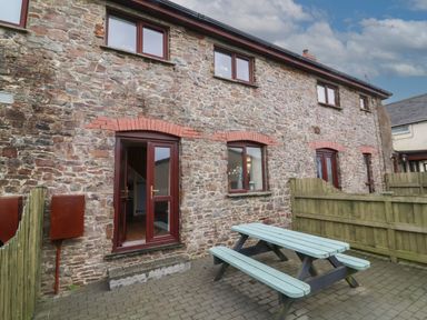 An outdoor patio area with a wooden picnic table beside a stone house with multiple windows and doors at Badgers Sett in Stibb near Kilkhampton