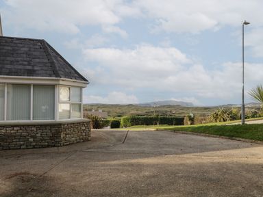 A driveway next to a house with stone wall and large windows overlooking a grassy landscape with hills and a street lamp at Horn Head View in Carrigart County Donegal