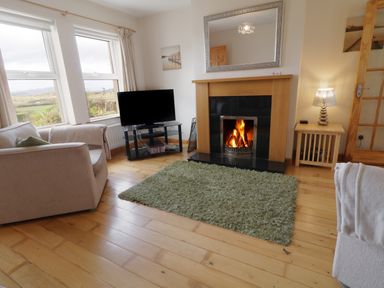 A living room with a fireplace a TV on a stand two beige sofas a small wooden side table with a lamp and a green rug on a wooden floor at Horn Head View in Carrigart County Donegal