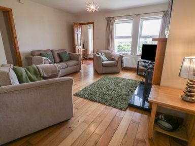 A living room with beige sofas green cushions a green rug wooden floor a TV on a stand and a wooden side table with a lamp at Horn Head View in Carrigart County Donegal