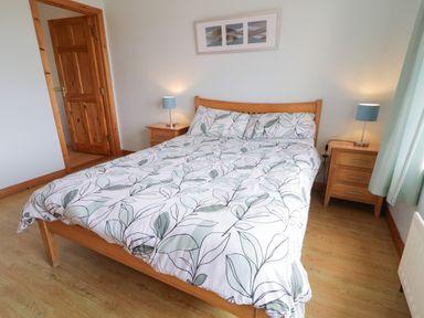 A bedroom with a wooden double bed white and green leaf patterned bedding two wooden bedside tables with blue lamps a wooden door and a window with light curtains at Horn Head View in Carrigart County Donegal