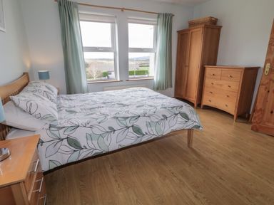 A bedroom with a bed, wooden wardrobe, chest of drawers, bedside table, and two windows with curtains at Horn Head View in Carrigart County Donegal