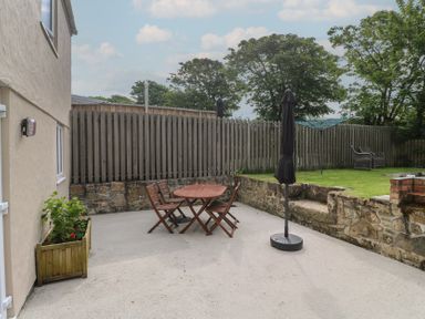 A backyard patio with a wooden table and chairs a closed umbrella a planter box and a grass area with a wooden bench at The Packing House in Rosudgeon