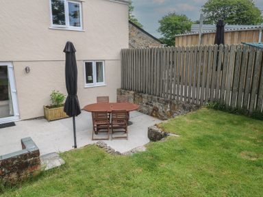 A backyard patio with a wooden table and chairs an umbrella closed beside a cream house with a grassy area and wooden fence at The Packing House in Rosudgeon