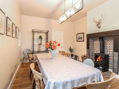 A dining room with a long table covered with a tablecloth and a vase with flowers a fireplace with a wood burning stove and decorative tiles and several chairs around the table at Grosvenor House in Kirkley Rise