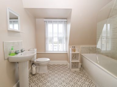 A bathroom with a sink, toilet, window with blinds, patterned floor, bathtub with glass screen, and a white shelving unit at Grosvenor House in Kirkley Rise