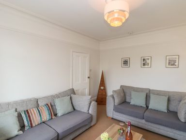 A living room with two grey sofas and cushions a wooden coffee table with flowers and a bottle three pictures on the wall and a wooden cone shaped cabinet at Trackside Ruswarp