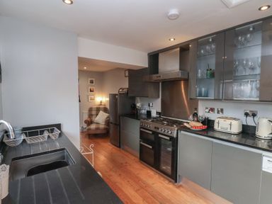 A kitchen with black countertops gray cabinets a stove a toaster and a kettle with a chair and framed pictures in the background at Trackside in Ruswarp